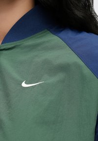 Πράσινο και μπλε αθλητικό μπουφάν με ριμπ γιακά, με λευκό λογότυπο Nike ραμμένο στην περιοχή του στήθους. Ορατή υφή υφάσματος.