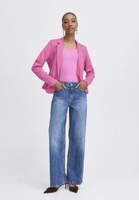 Rosa Blazer über einem rosa Tanktop; blaue Jeans mit weitem Bein; braune Absatzschuhe. Model steht mit Händen in der Hüfte vor einem schlichten Hintergrund.