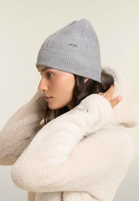 Beanie grigio a maglia con risvolto a coste e dettaglio del logo, indossato con una morbida felpa in pile color crema. Focus su abbigliamento invernale funzionale.