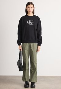 Čierna mikina s bielym logom, olivovozelené široké nohavice a čierne robustné čižmy. Zobrazuje minimalistický dizajn a textúrovanú tkaninu.