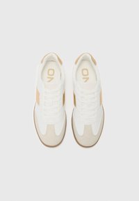 White sneakers with beige accents, featuring a textured suede toe, lace-up front, and branding on the tongue. Rubber sole with patterned grip.