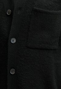 Melnas vilnas adītas kardigāns ar trim melnajām pogām un kvadrātveida krūšu kabatu uz teksturētas auduma.