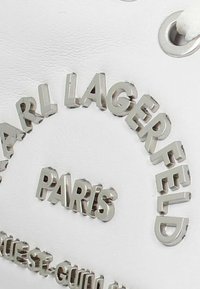 Vit sneaker med upphöjd silverbokstäver, med "CARL LAGERFELD" och "PARIS" präglat på sidan. Slät och texturerad yta.