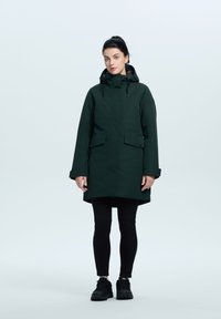 Parka impermeable verde oscuro con capucha, cierre frontal y bolsillos con botón a presión. Textura suave y corte recto. Llevado sobre leggings negros y zapatos.