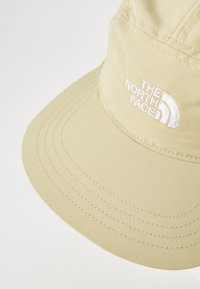 Beige cap with a wide brim, made of smooth fabric. Features white embroidered logo on the front and detailed stitching along the brim.