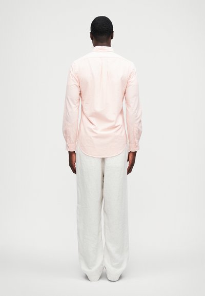 Koszula z długim rękawem w pastelowym różowym kolorze w pionowe paski, zestawiona z luźnymi, białymi spodniami z lnu, widok z tyłu.