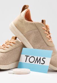 Beige Leinenschuhe mit orangenen Schnürsenkeln, strukturiertem Stoff und leicht gesprenkelten Sohlen. Einschließlich einer blauen Karte mit "TOMS" in fettgedruckten schwarzen Buchstaben.
