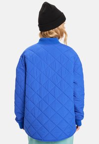 Modrá prošívaná bunda s diamantovým vzorem, žebrovaným límcem a manžetami. Měkká textura a volný střih; nošena s černou čepicí.
