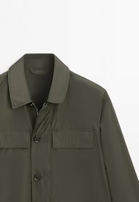 Giacca a maniche lunghe verde scuro con colletto appuntito, due tasche a patta sul petto, chiusura frontale con bottoni e una cerniera visibile sotto il pattino.