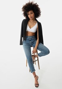 Zwarte blazer over een witte bralette, hoge lichtblauwe spijkerbroek en zwarte sandalen met bandjes. Krullend haar; zittend op een houten kruk.