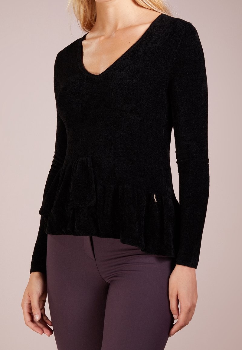 Femme portant un haut noir à manches longues avec un col en V et un ourlet à volants, associé à un pantalon ajusté violet foncé, visible des épaules aux hanches.