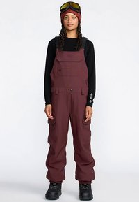 Maroon snow overalls with adjustable straps, zippered pockets, and a loose fit. Paired with a black long-sleeve top and goggles.