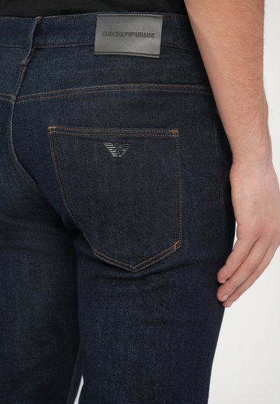 Sötétkék farmernadrág rézszínű varrással, fekete bőr derékpánttal és ezüst logó díszítéssel a hátsó zsebnél.