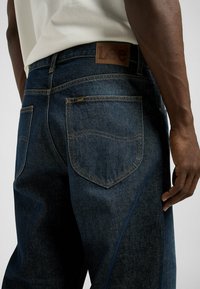 Dark blue denim jeans featuring a leather patch with "Lee" branding, visible stitching, and five-pocket design. Texture appears smooth and durable.