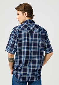 Camicia a maniche corte con un motivo a quadri blu e navale, colletto button-down e taglio dritto. Il tessuto sembra leggero e con una texture.