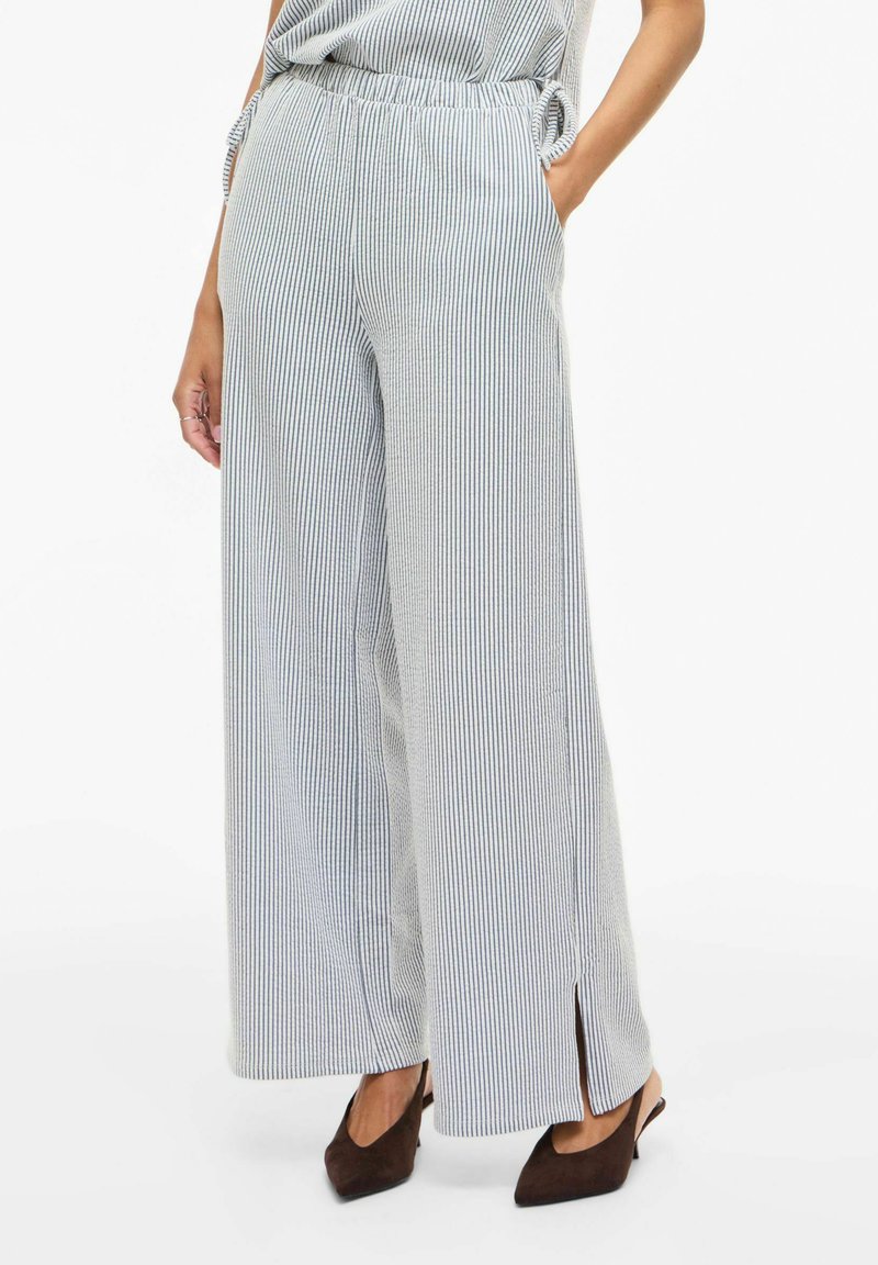 Femme portant un pantalon rayé à jambes larges et taille haute avec des fentes latérales, ainsi que des escarpins marron à bouts pointus, debout avec une main dans la poche.