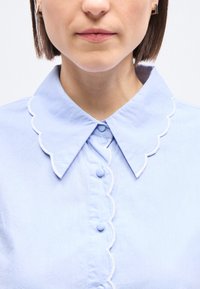 Γυναίκα που φοράει ανοιχτό γαλάζιο πουκάμισο με κυματιστές άκρες στο γιακά και στη σειρά με τα κουμπιά, δείχνοντας το κάτω μέρος του προσώπου και το λαιμό της.