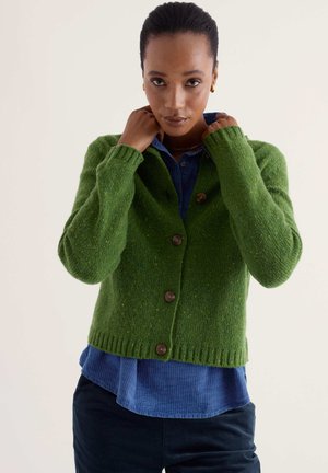 Femme ajustant le col d’un cardigan vert boutonné porté sur une chemise bleue et un pantalon foncé sur un fond neutre.