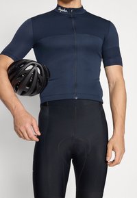 Camiseta de ciclismo azul marino con cremallera y mangas cortas, combinada con pantalones cortos acolchados negros. Un casco negro se sostiene en la mano izquierda.
