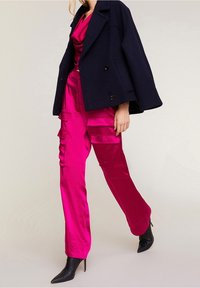 Manteau peacoat coupé marine sur pantalon cargo fuchsia en satin avec plusieurs poches, associé à des escarpins noirs à bout pointu.