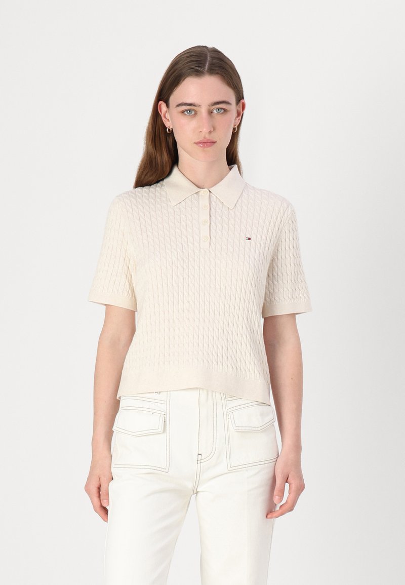 Kortermet beige polo-skjorte med teksturert strikkedesign, kragehals og lite logo på brystet. Kombinert med lyse bukser.