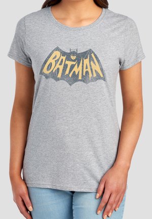 Frau trägt ein graues T-Shirt mit einem gelb-schwarzen Batman-Logo und Schriftzug auf der Brust, kombiniert mit hellblauen Jeans.