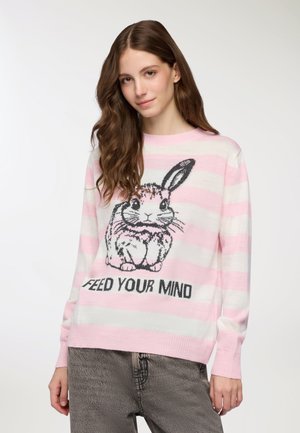 Giovane donna con i lunghi capelli castani indossa un maglione a righe rosa e bianche con un'illustrazione di un coniglio e il testo "NUTRI LA TUA MENTE."