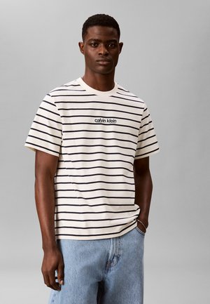 Junger Mann steht mit einer Hand in der Tasche, trägt ein weiß-schwarz gestreiftes Calvin Klein T-Shirt und hellblaue Jeans.