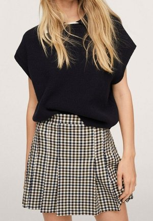 Kvinde iført en sort ærmeløs striktop og en høj talje plisseret mininederdel med sort, hvid og beige ternet mønster.