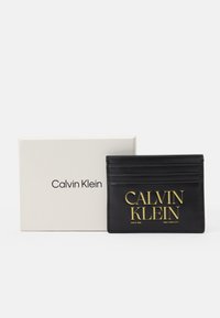 Černý kožený držák na karty s více přihrádkami a zlatě embosovaným logem "CALVIN KLEIN". Dodává se v bílé značkové krabičce.