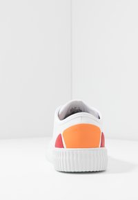 Sneaker bianca con suola in gomma texturizzata, forma arrotondata e accenti colorati in arancione e rosso nell'area del tallone.