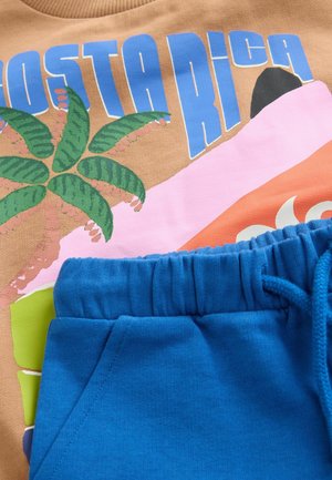 Brauner Sweatshirt mit Grafik "COSTA RICA", das einfarbige Palmen und ein abstraktes Design zeigt. Blaue Shorts mit elastischem Bund und Taschen.