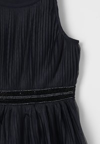 Mouwloze zwarte geplooide jurk met een ronde halslijn en een pailletten tailleband detail op een witte achtergrond.