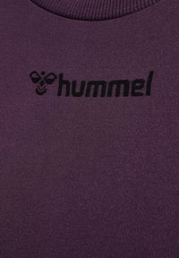 Lila sportskjorta med ribbad krage, som har en svart logotyp som säger "hummel." Tyget har en slät, något glänsande yta.