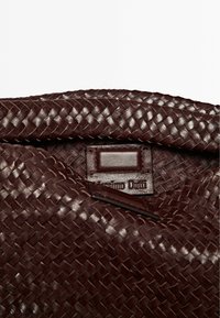 Borsa a tracolla in pelle intrecciata marrone scuro con una texture liscia, dotata di un'etichetta rettangolare con il nome del marchio e una chiusura a patta superiore.