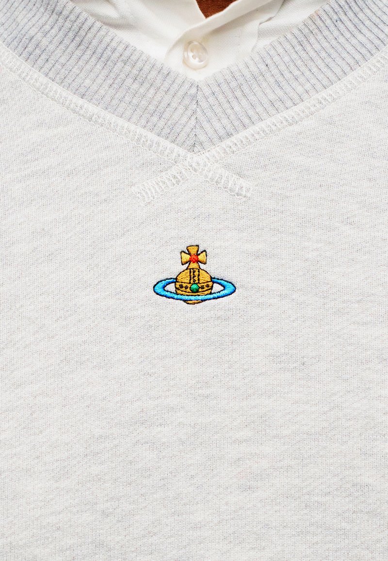 Maglione grigio chiaro in maglia con camicia bianca colletto sotto, con piccolo ricamo di un globo dorato con un anello blu e una croce rossa sul petto.