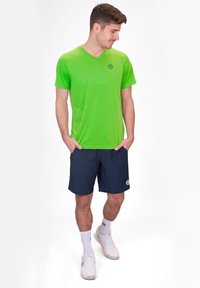 Camiseta verde de manga corta y cuello en V, hecha de un material ligero; combinada con pantalones cortos azul marino, zapatillas blancas y calcetines tobilleros.