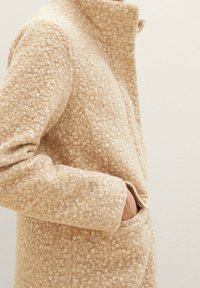 Beige ullblandad kappa med en texturerad yta, hög krage och knappstängning. Har sidofickor och en lös passform.