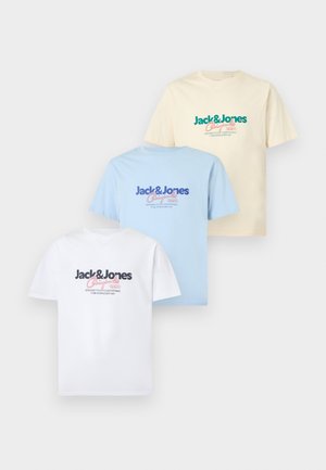 Νέος άντρας φοράει λευκή μπλούζα με λογότυπο Jack & Jones, μαύρο παντελόνι και ασημένια αθλητικά παπούτσια, στέκεται με το ένα χέρι στην τσέπη.