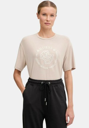Donna che indossa una t-shirt beige Guess con logo rotondo e pantaloni neri con coulisse, in piedi con una mano in tasca.