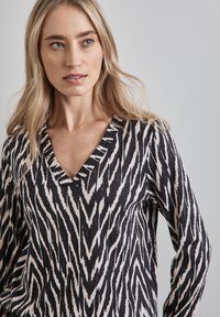 Czarna i biała bluzka w wzory, z głębokim dekoltem w kształcie litery V i długimi rękawami. Materiał ma teksturowany wygląd z wzorem w zygzak.