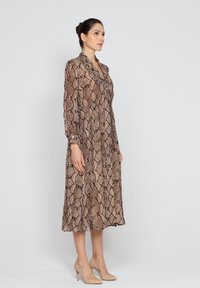 Brązowa sukienka w nadruk imitujący skórę węża, z długimi rękawami i kołnierzykiem, sięgająca do połowy łydki. Noszona z beżowymi szpilkami.