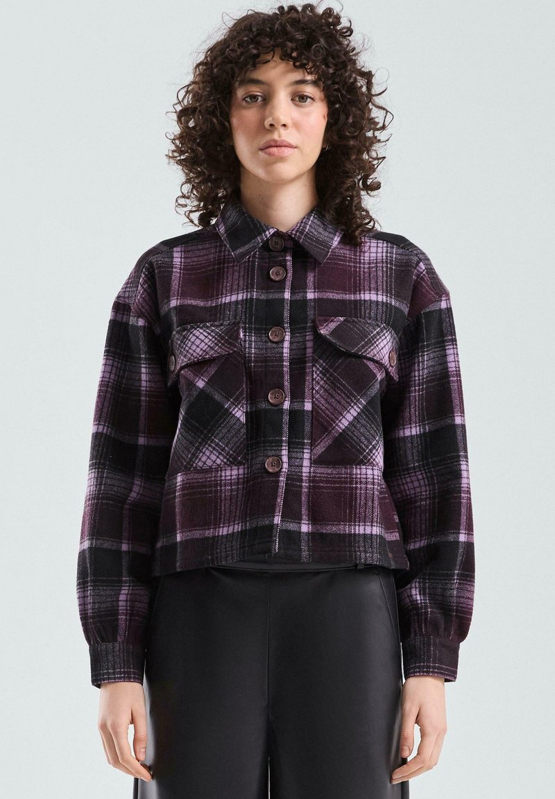 Fioletowo-czarna flanelowa kurtka w kratę z zapięciem na guziki, dwoma kieszeniami na piersi i długimi rękawami. Materiał o fakturze, krótki dół.