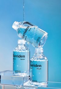Klare glasflasker med Torriden serum, der har lyseblå etiketter med sort tekst. Nogle flasker står oprejst, en er skrå, med dråber synlige.