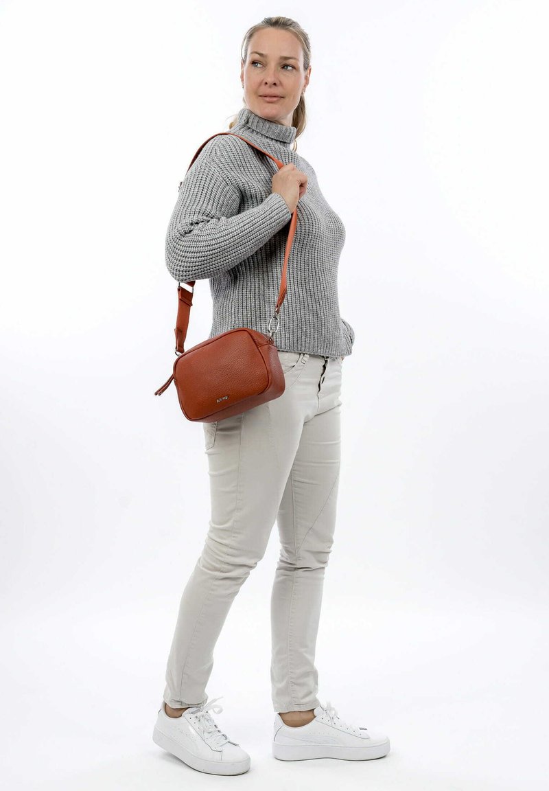 Brown leather crossbody bag with an adjustable strap, zip closure, rectangular shape, and textured surface. Worn with a gray sweater and beige pants.
