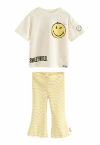 Crème kortgeschouderd t-shirt met zwarte tekst "SMILEYWORLD" en een gele smiley-afbeelding, gecombineerd met gele geruite flared leggings.