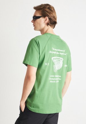 Tricou verde din bumbac, cu un grafism al unui coș de baschet și text pe spate. Mâneci scurte și un croi relaxat.