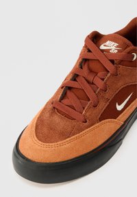 Nike SB apavi ar apdedzinātu oranžu zamšu augšdaļu, ar sieta akcentiem, plakano auklu un melnu gumijas zoli; iekļauj balto Nike swoosh simbolu.