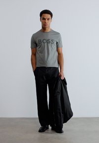 Νεαρός άνδρας στέκεται μπροστά σε απλό τοίχο φορώντας γκρι μπλουζάκι BOSS και μαύρο παντελόνι, κρατώντας μαύρο σακάκι σε ένα χέρι.