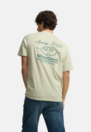 Joven con camiseta beige claro con raquetas de tenis y texto "Silbon Classic" en la parte trasera, combinado con jeans azules.